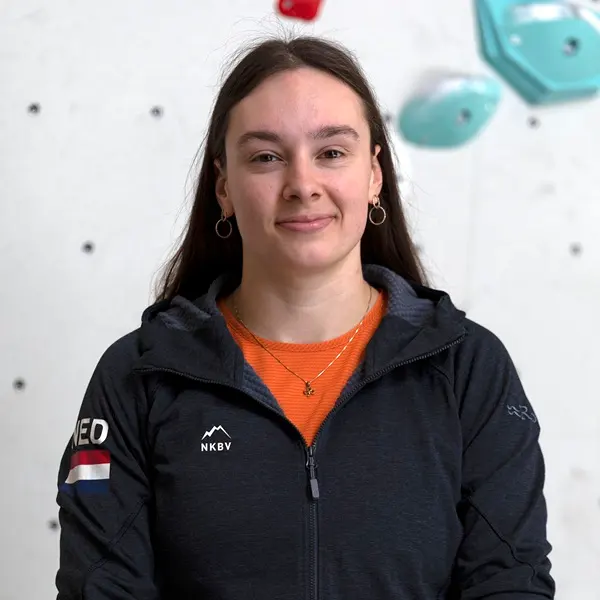 Een jonge vrouw met lang bruin haar, gekleed in een donker jasje met een patch in de vorm van de Nederlandse vlag, een oranje shirt en gouden hoepeloorbellen, staat binnenshuis voor een klimwand met b
